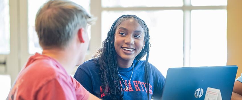 Two students sitting at table with laptop talking.